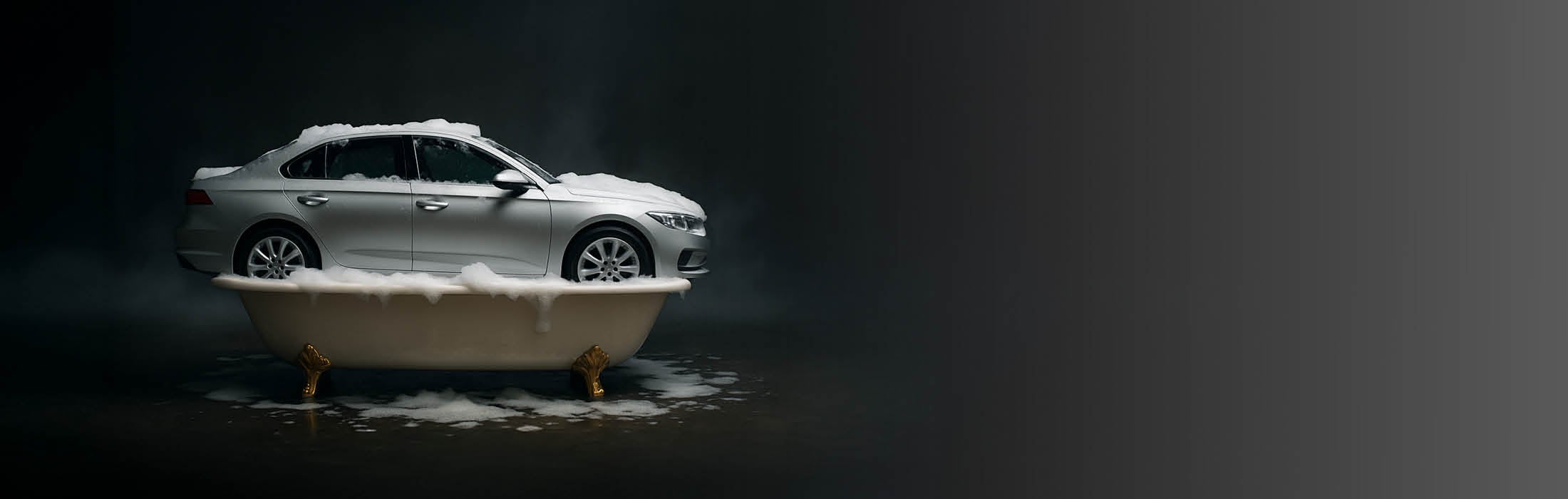 Auf dem Bild ist ein silbernes Auto zu sehen, das in einer klassischen weißen Badewanne mit goldenen Füßen steht. 